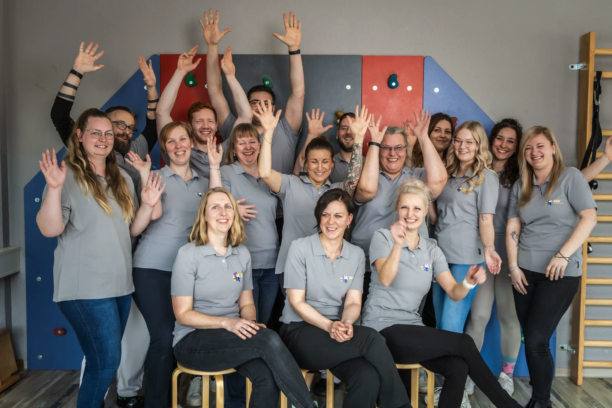 Das Team der 360° Therapie Kesselsdorf winkt vor der Kletterwand im Kursraum